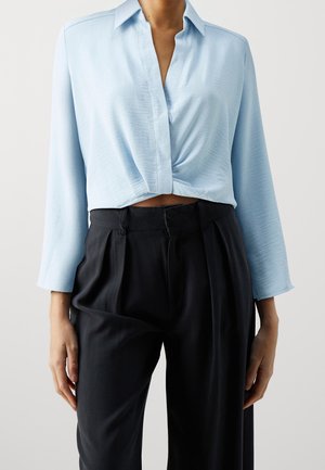 Blouse bleu clair à manches longues avec col et devant torsadé, associée à un pantalon noir plissé taille haute.