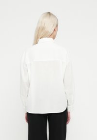 Bela srajca z dolgimi rokavi in sproščenim krojem, z obdanim robom in gumbi na manšetah. Na hrbtu je šiv preko ramen za dodatno podrobnost.