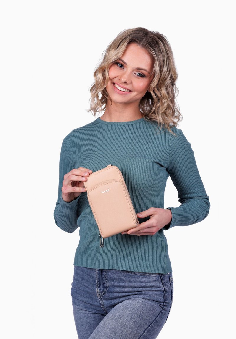 Přebal v broskvové barvě se zipem, hladkým povrchem, přední kapsou a jemným logem, který drží osoba v tyrkysovém žebrovaném svetru.