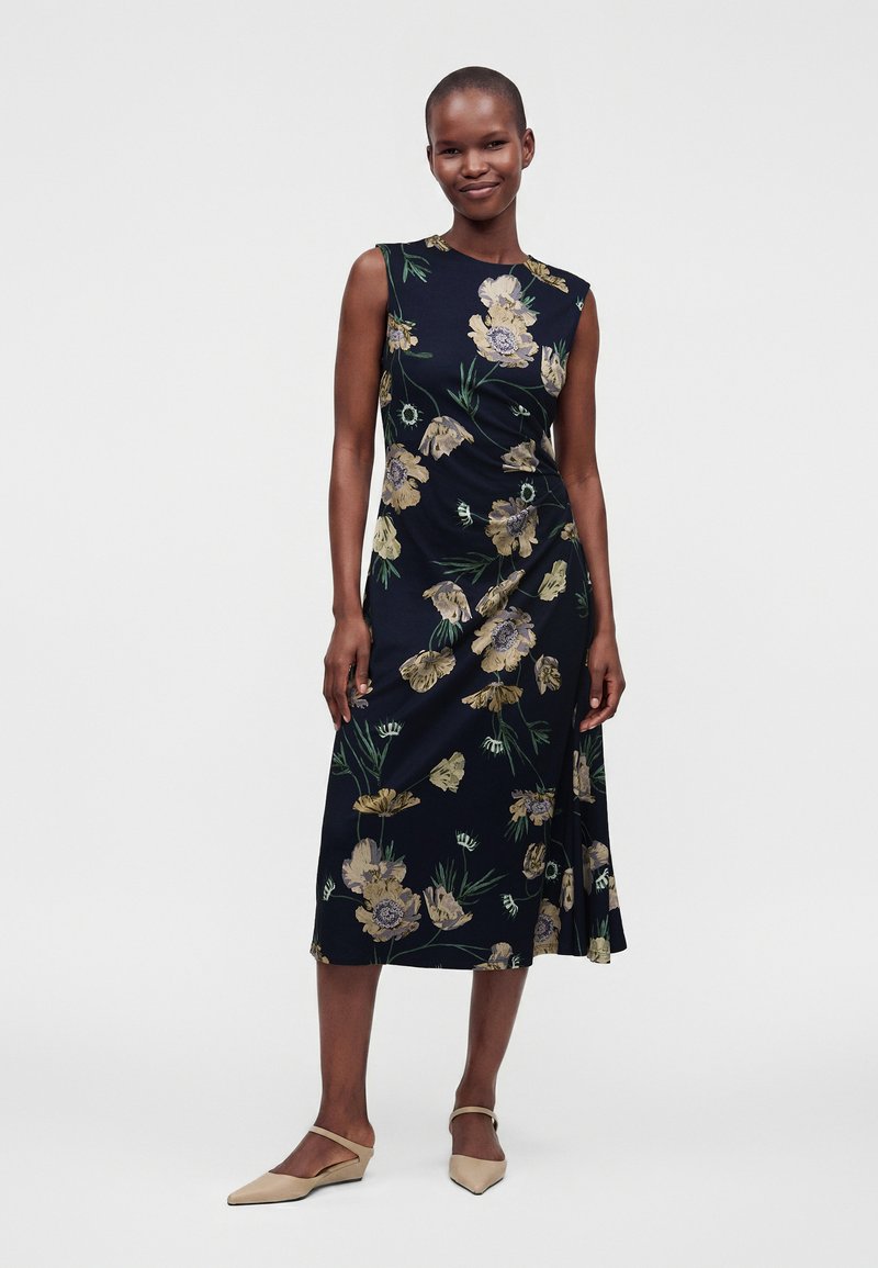 Woman standing and smiling, wearing a sleeveless black dress with a yellow floral pattern and beige flat shoes against a plain white background.