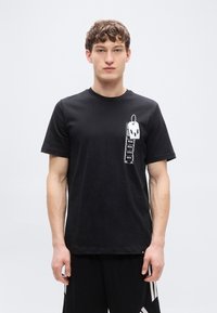 Μαύρο βαμβακερό t-shirt με εκτυπωμένο γραφικό από ετικέτες αποσκευών με κείμενα στην αριστερή πλευρά. Κοντά μανίκια και στρογγυλή λαιμόκοψη.