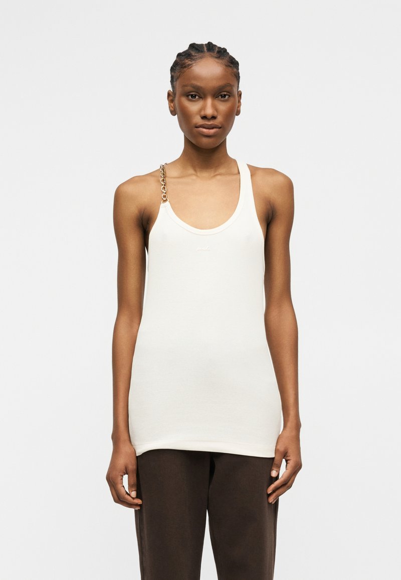 Γυναίκα με πλεξούδες στα μαλλιά, που φορά λευκό αμάνικο τοπ με χρυσή αλυσίδα ως λουρί στον έναν ώμο και σκούρο καφέ παντελόνι, στέκεται μπροστά σε απλό φόντο.