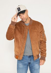 Bruine corduroy bomberjack met ribgebreide manchetten en kraag, voorzien van zijzakken en een ritszak op de mouw. Draagt een grijs shirt en pet.