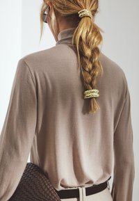 Pitkähiha, vaaleanruskea neulepaita, jossa on korkea kaulus. Siinä on punos, joka on kiinnitetty kultaisella hiuslisäosalla. Kudottu ruskea laukku etualalla.