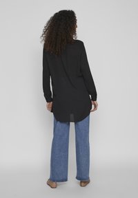 Chemise noire à manches longues avec un ourlet incurvé, fabriquée en tissu lisse, associée à un jean bleu à jambes larges et des chaussures plates marron.