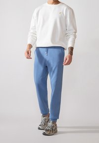 Biała bawełniana bluza z długim rękawem, w połączeniu z niebieskimi spodniami dresowymi z elastycznym paskiem w talii i srebrnymi sznurówkami. Szare i srebrne buty sportowe.