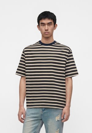 Jeune homme debout face à l'avant, portant un t-shirt à manches courtes rayé bleu marine, blanc et beige, et un jean bleu clair déchiré.