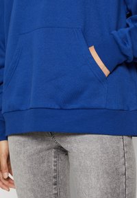 Niebieska bawełniana bluza z kapturem z przednią kieszenią. Prążkowany dół i mankiety. W połączeniu z jasnoszarymi dżinsami. Gładka tekstura, casualowy design.