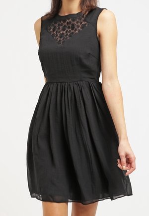 Robe noire sans manches avec une taille et une jupe ajustées. Le corsage présente un panneau en dentelle florale transparente. Le tissu texturé ajoute de la profondeur.