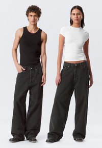 Due modelli indossano jeans in denim nero larghi. L'uomo ha una canotta nera senza maniche, mentre la donna indossa una maglietta aderente bianca a maniche corte.
