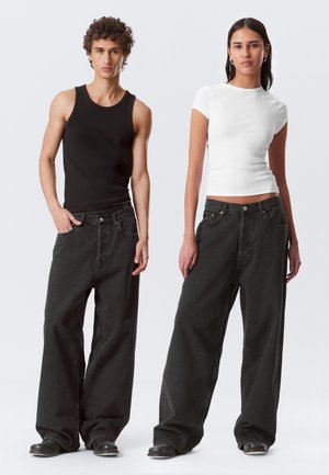 Schwarzes Tanktop und weißes kurzärmliges Hemd, getragen mit weit geschnittenen, dunklen Jeans. Die Jeans verfügen über Fronttaschen und einen Knopfverschluss, beide Modelle tragen schwarze Schuhe.