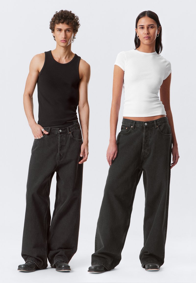 Due modelli indossano jeans in denim nero larghi. L'uomo ha una canotta nera senza maniche, mentre la donna indossa una maglietta aderente bianca a maniche corte.