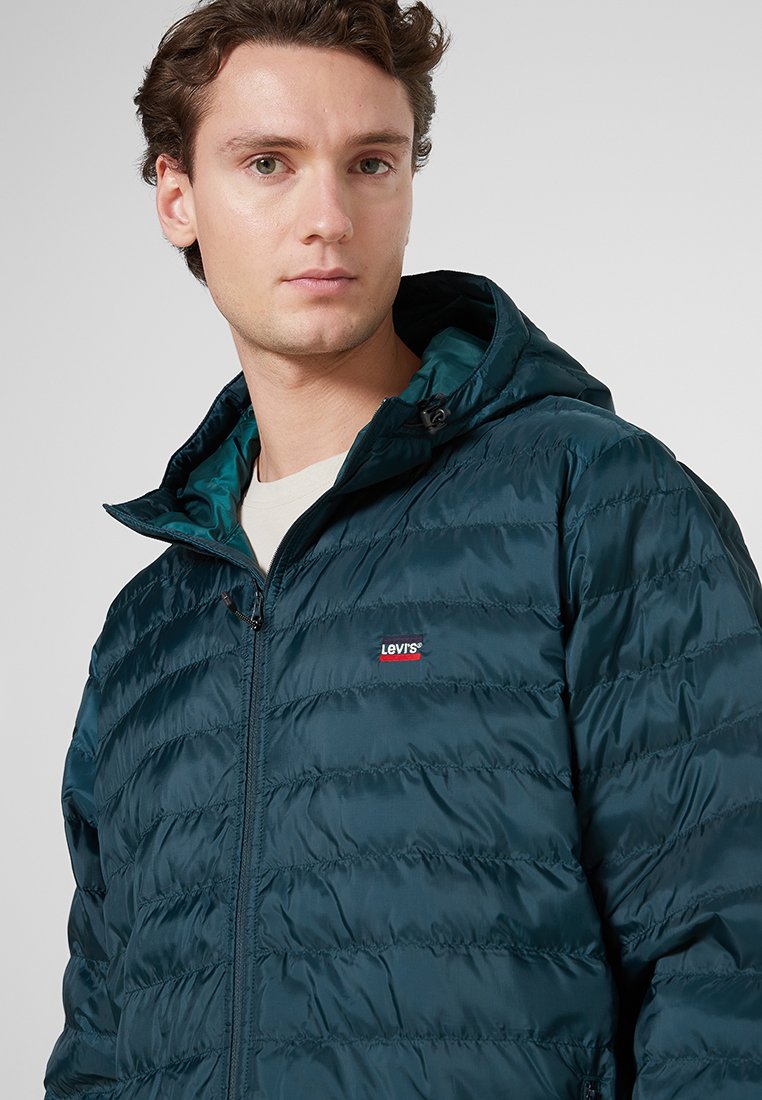 Donker teal pufferjack met kap, met een ritssluiting aan de voorkant, een gequiltde textuur en een klein Levi's logo op de borst. Voering aan de binnenkant.