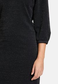 Musta mekko, jossa on teksturoitu, kimaltava pinta. Varustettu kolme neljännestä hihasta ja rullattua hihansuuta ranteen lähellä.