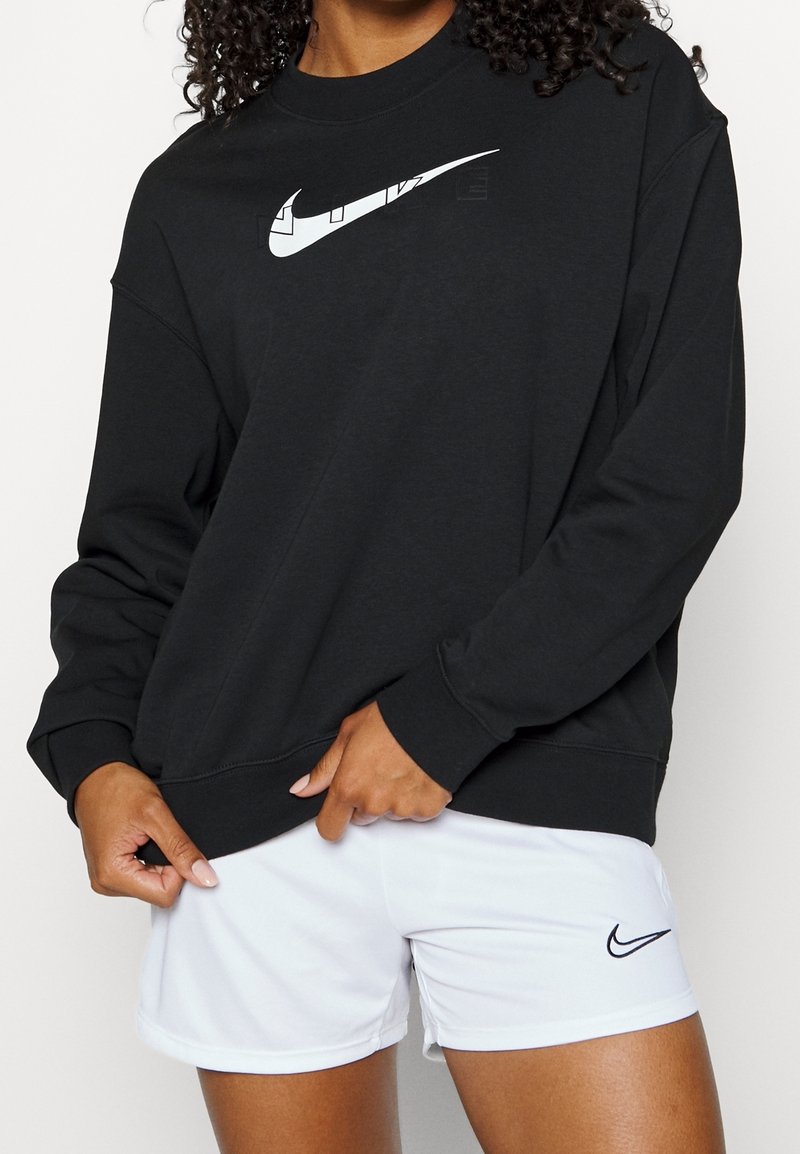 Personne portant un sweat-shirt Nike noir avec un logo blanc et un short Nike blanc tirant légèrement l'ourlet du sweat-shirt vers le bas.