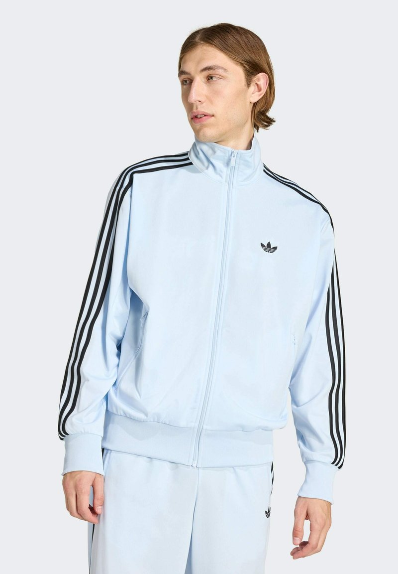 Giovane uomo che indossa una tuta Adidas azzurro chiaro con strisce nere sulle maniche e sui pantaloni, in piedi contro uno sfondo grigio.