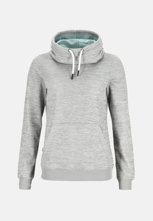 Kobieta stoi z rękami w kieszeniach, ubrana w dopasowany jasnoszary hoodie i dresy, z białymi sneakersami na białym tle.