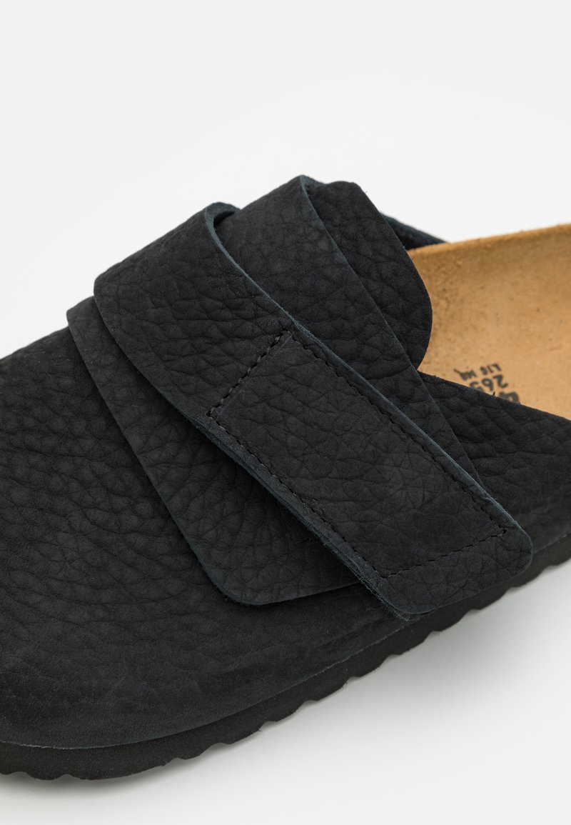 Chaussure noire à enfiler avec une surface texturée, dotée d'une large sangle Velcro. L'intérieur est beige et la semelle présente un motif ondulé.