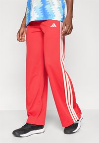 Osoba w czerwonych spodniach dresowych Adidas z białymi paskami i czarnych sneakersach, stojąca na jednolitym jasnym tle.