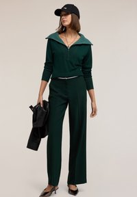 Donkergroen gebreide pullover met een brede kraag, gecombineerd met rechttoe rechtaan broek. Zwarte pet en glanzende zwarte schoenen met spitse neus, met een zwart jack in de hand.