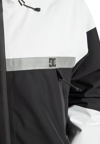 Zwart-witte jas met een glanzende zilveren band en DC-logo. Heeft een ritssluiting en een verstelbare trekkoord bij de kraag.