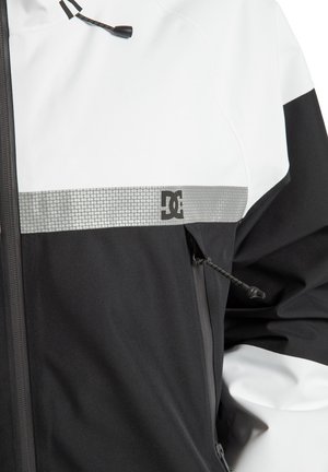 Schwarz-weiße Jacke mit einem glänzenden silbernen Streifen und DC-Logo. Verfügt über einen Reißverschluss und eine verstellbare Schnürung am Kragen.
