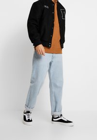 Černá varsity bunda s bílými detaily, hnědá mikina pod ní, světle modré zahnuté džíny a černobílé tenisky.