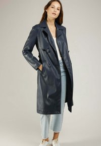 Manteau trench en cuir marine avec de grands revers, boutons double boutonnage, deux poches avant et une coupe longueur genou. Associé à un pantalon en denim clair.