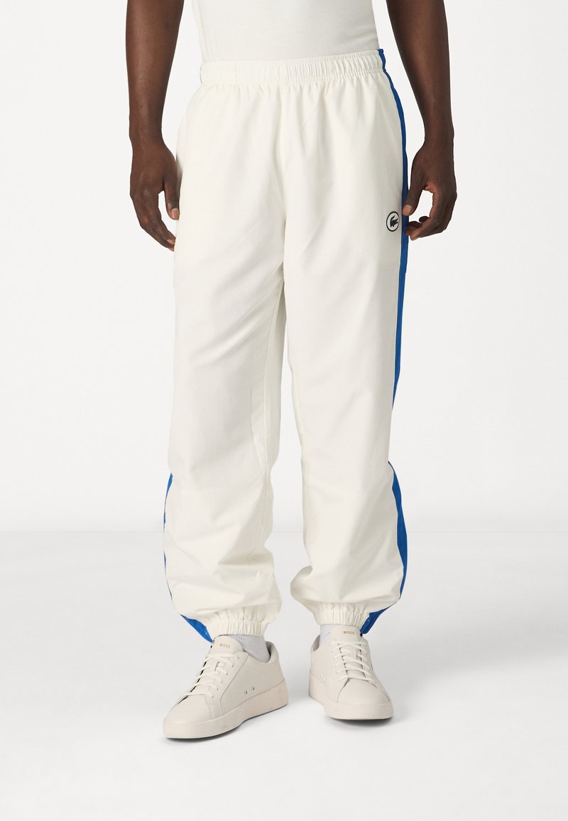 Pantalons de sport blancs avec des bandes latérales bleues, taille élastique et poignets. Associés à des baskets de couleur claire au design minimaliste.