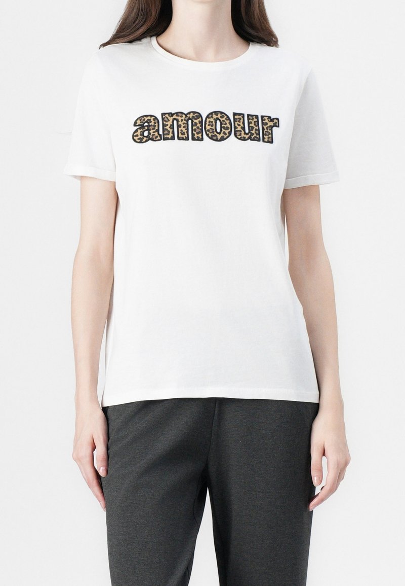 T-shirt blanc à manches courtes avec le mot "amour" en police imprimé léopard porté par une personne portant un pantalon foncé.