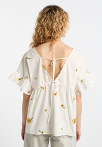 Blusa bianca a maniche corte con ricami floreali gialli e schiena profonda a V, dotata di chiusura a fiocco e maniche arricciate. Tessuto in cotone morbido.