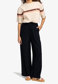 Jasno-beżowa koszula z krótkim rękawem w poziome brązowe i koralowe paski, zestawiona z luźnymi czarnymi spodniami i jasnobrązowymi sandałami.