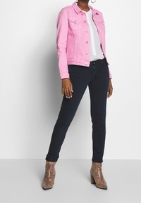 Chaqueta de mezclilla rosa con cierre de botones, combinada con camisa blanca, jeans oscuros y botines de tacón marrones con estampado de serpiente. Diseño sencillo y ajustado.