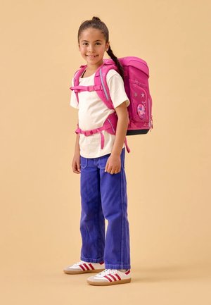 Pink backpack with reflective straps, star pattern, and a character patch. Paired with a white shirt, blue jeans, and striped sneakers.