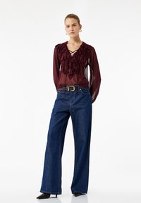 Blusa trasparente color bordeaux con dettagli di rouches e laccio, abbinata a jeans in denim blu a vita alta e gamba larga, con tacchi neri.