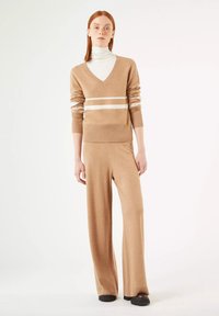 Beiger Strickpullover mit V-Ausschnitt und weißen Streifen, kombiniert mit passenden weit geschnittenen Hosen, die Rippbündchen und -saum haben. Schwarze Schuhe.