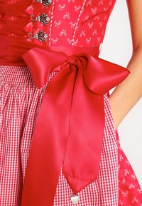 Robe rouge avec un corsage ajusté agrémenté de motifs floraux et de détails en dentelle. La jupe présente un motif à carreaux, maintenu par un grand nœud en satin.