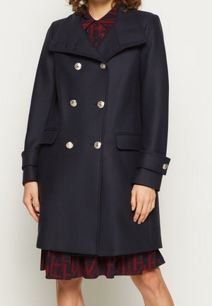Frau mit einem dunkelmarineblauen, zweireihigen Mantel mit silbernen Knöpfen über einem rot-schwarz gemusterten Kleid, stehend vor einem einfarbigen Hintergrund.
