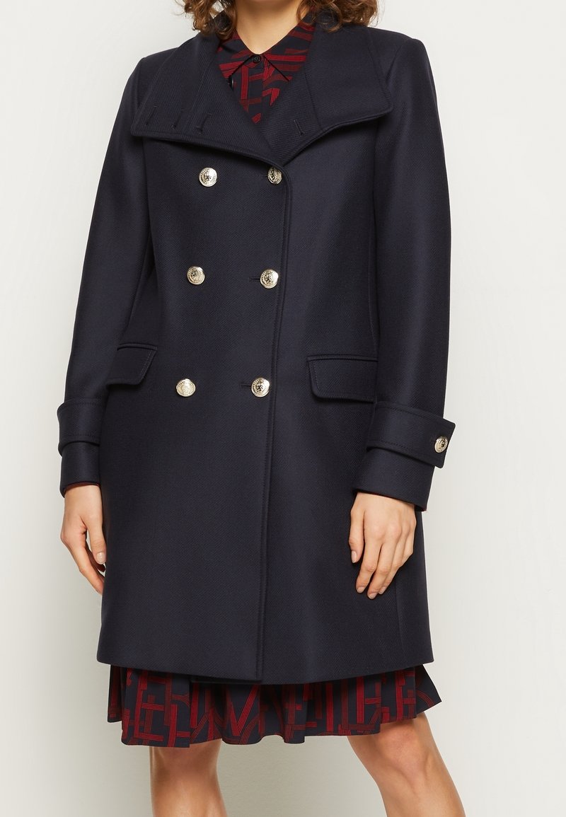 Femme portant un manteau croisé bleu marine foncé avec des boutons argentés sur une robe rouge et noire à motifs, debout devant un fond uni.