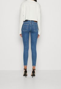 Mujer con jeans ajustados azul, sandalias de tacón negro y una blusa blanca de manga larga y cropped, de pie frente a un fondo simple.