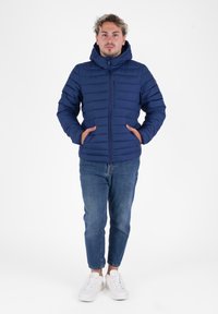 Jonge man staat met zijn handen in zijn zakken, gekleed in een blauwe gewatteerde jas, blauwe spijkerbroek en witte sneakers op een witte achtergrond.