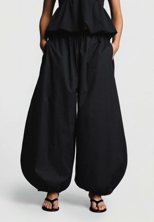 Pantalon large noir avec taille élastique et ourlets froncés, porté avec un haut noir et des sandales noires, mains dans les poches, debout sur un fond uni.