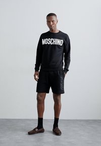 Sudadera negra con "MOSCHINO" en letras blancas; combinada con pantalones cortos negros que presentan una marca en relieve. Modelo con zapatos oscuros y calcetines.