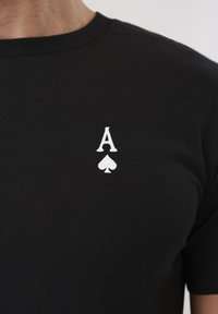 Μαύρο μπλουζάκι με λευκό γράμμα Άσσο και σύμβολο σπαθί τυπωμένα στο πάνω αριστερό μέρος του στήθους.