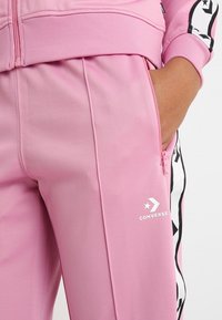 Rosa Jogginghosen mit Reißverschlusstaschen, die eine glatte Textur, das Converse-Logo und schwarze Streifen an den Seiten aufweisen.