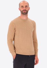 Uomo con barba e capelli corti che indossa un maglione beige a maniche lunghe e pantaloni neri, in piedi con una mano in tasca contro uno sfondo semplice.