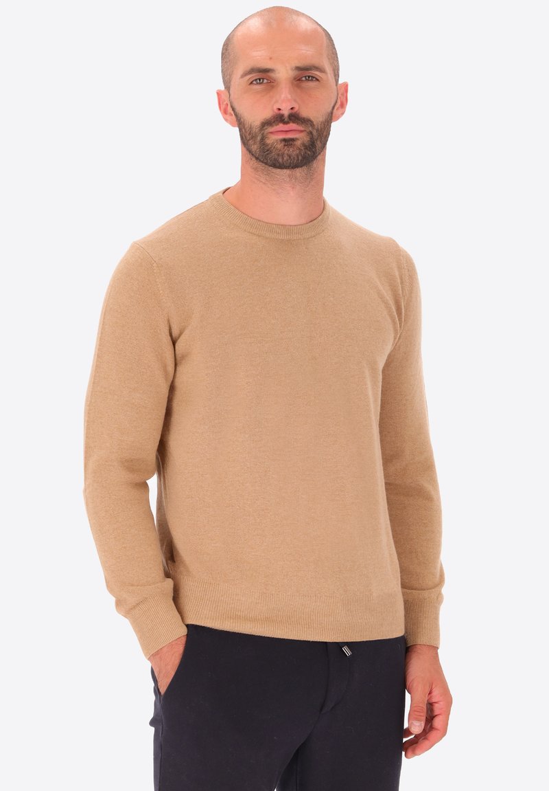 Uomo con barba e capelli corti che indossa un maglione beige a maniche lunghe e pantaloni neri, in piedi con una mano in tasca contro uno sfondo semplice.