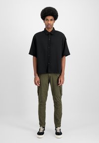 Melna īsām piedurknēm poga krekls, olīvzaļi cargo bikses ar sānu kabatām un elastīgām gumijām, melni sporta apavi ar baltām zolem.