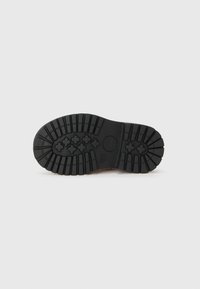 Semelle en caoutchouc noir d'une chaussure, présentant un motif de semelle texturé et un design arrondi, positionnée sur un fond blanc uni.
