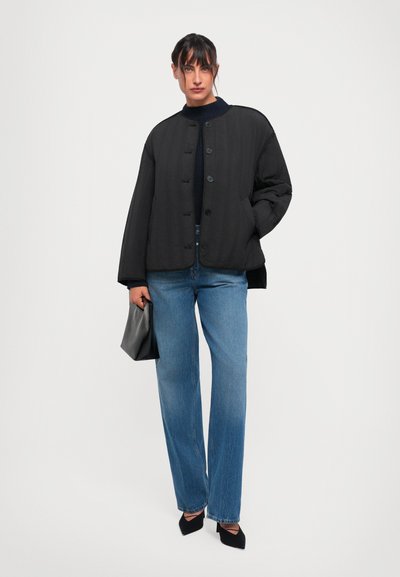 Frau steht in einem schwarzen, geknöpften Jacket, dunklem Pullover, blauen Jeans und schwarzen, spitzen Absätzen, hält eine schwarze Tasche vor einem weißen Hintergrund.